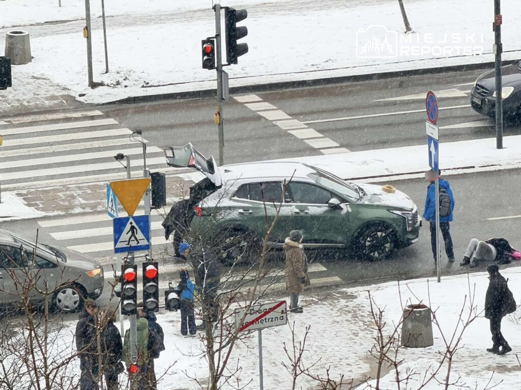 Zielony samochód stoi na przejściu dla pieszych, a obok dwóch mężczyzn rozmawia, podczas gdy śnieg pada na ulicę.