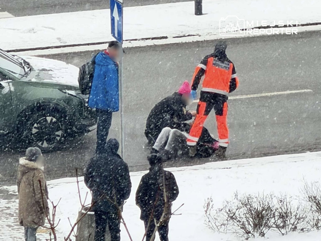 Zespół ratunkowy udziela pomocy osobie leżącej na ulicy w trakcie opadów śniegu. W tle widać samochód i przechodniów.