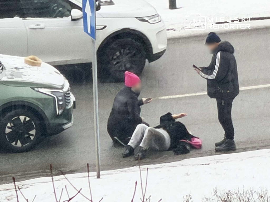 Dwie osoby siedzą na jezdni, podczas gdy jedna osoba stoi obok, trzymając telefon. W tle widać samochody na śniegu.