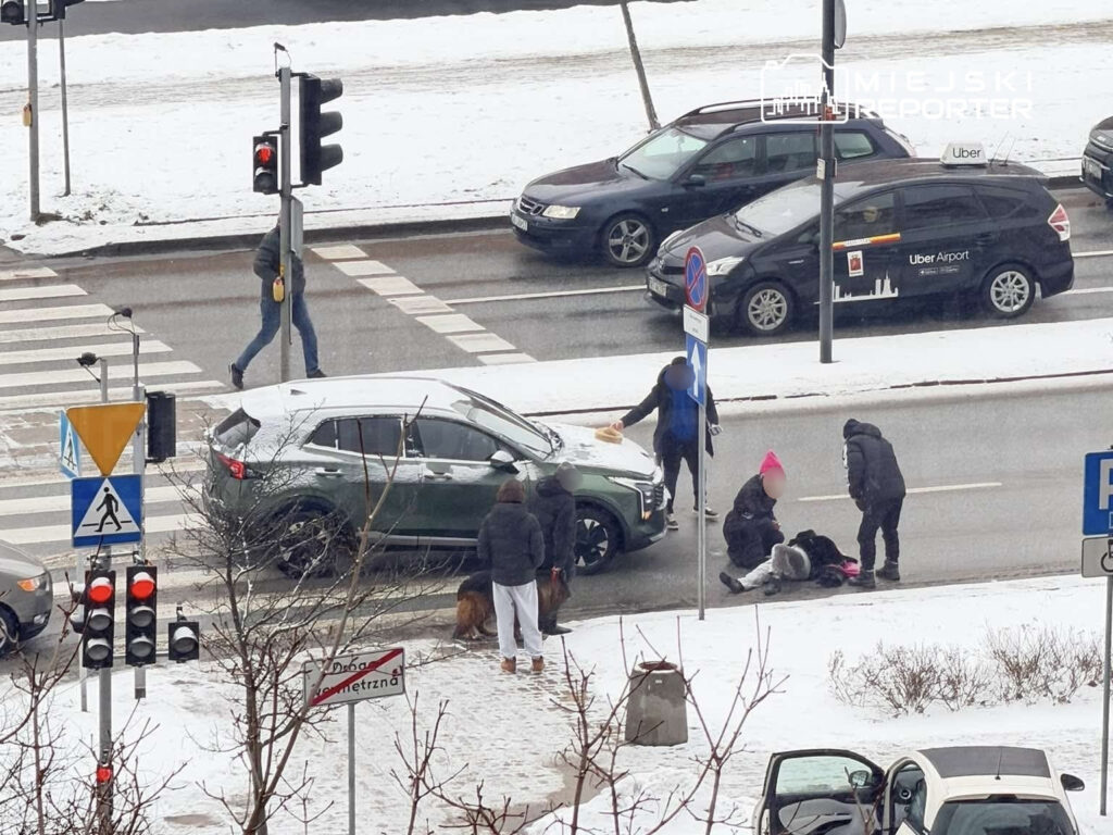 Grupa osób pomaga leżącej kobiecie na zaśnieżonym przejściu dla pieszych, obok stoi zaparkowany samochód.