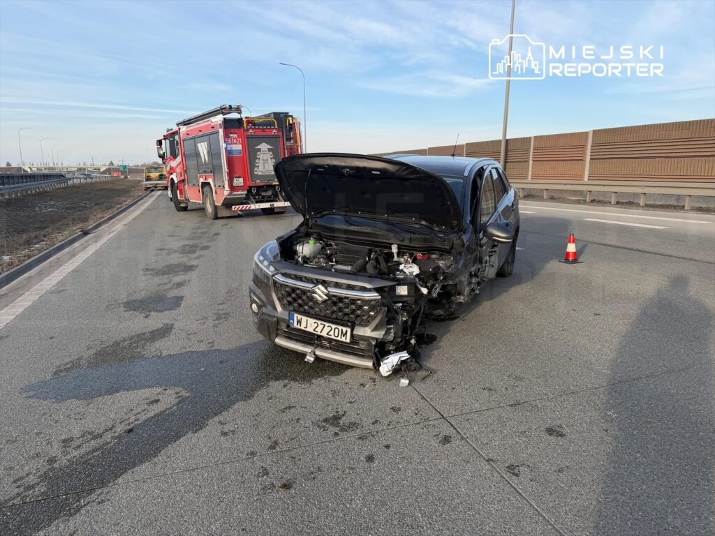 Uszkodzony czarny samochód osobowy stoi na poboczu drogi, obok wozu strażackiego. W tle widoczne są znaki drogowe.