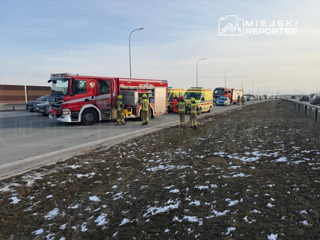 Strażacy w mundurach pracują przy wypadku drogowym na jezdni, obok pojazdów ratunkowych i zablokowanych samochodów.