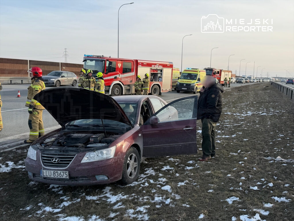 Mężczyzna w kurtce rozmawia przez telefon obok unieruchomionego samochodu, a w tle stoją strażacy i pojazdy ratunkowe.