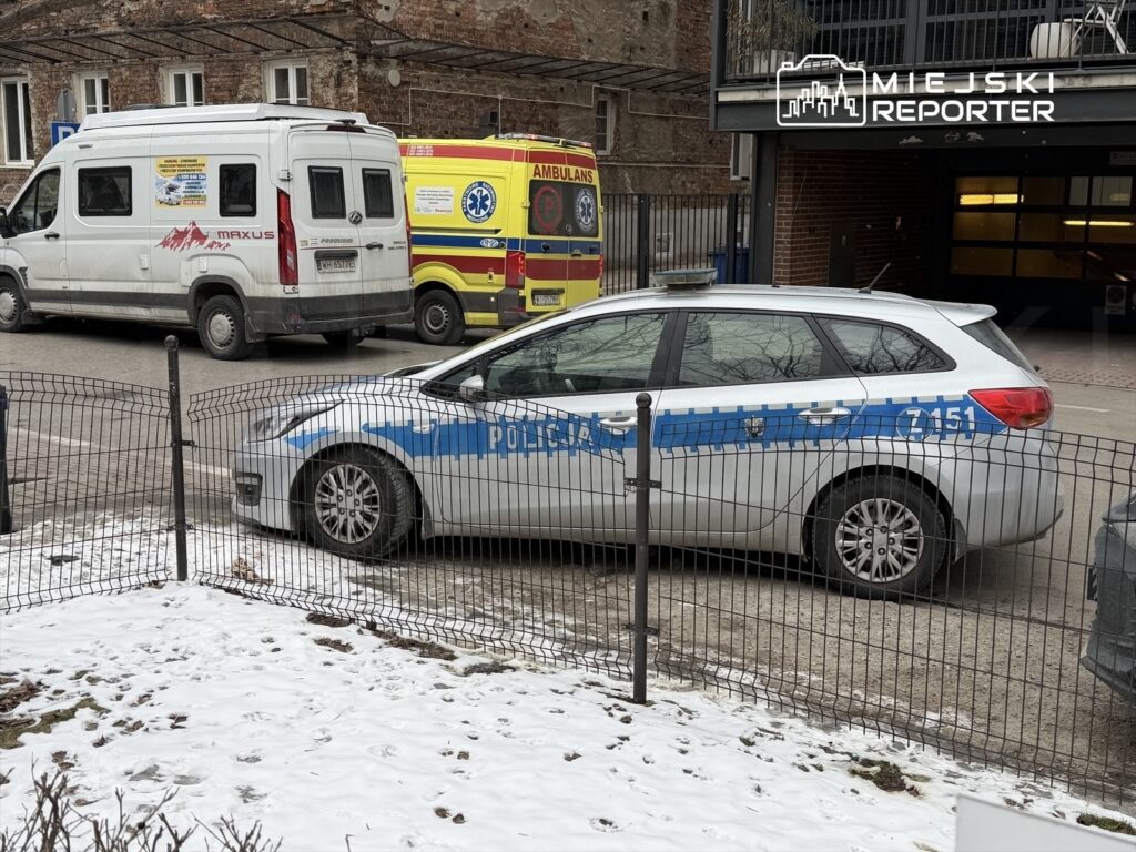 Policyjny radiowóz zaparkowany na śnieżnym podjeździe obok ambulansu i furgonetki w miejskim otoczeniu.