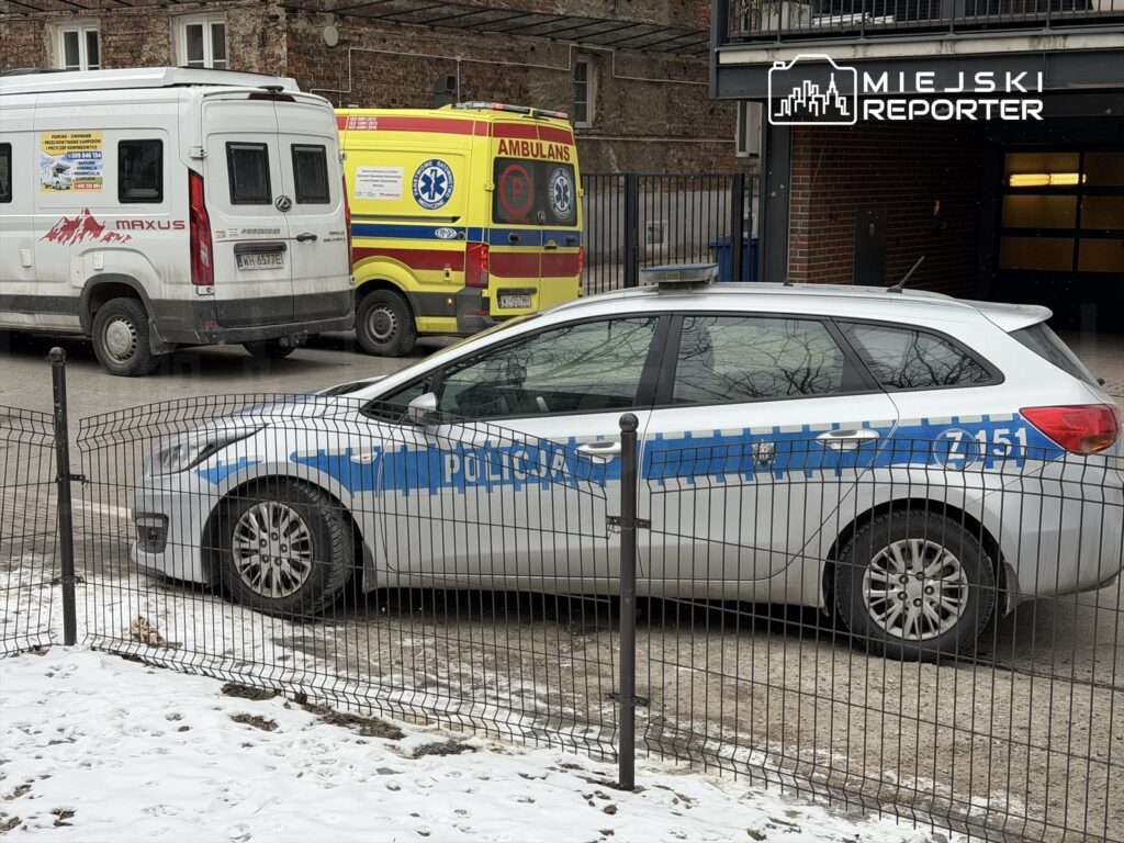 Radiowóz policyjny zaparkowany na ulicy obok ambulansu i dostawczego samochodu, w otoczeniu budynków.