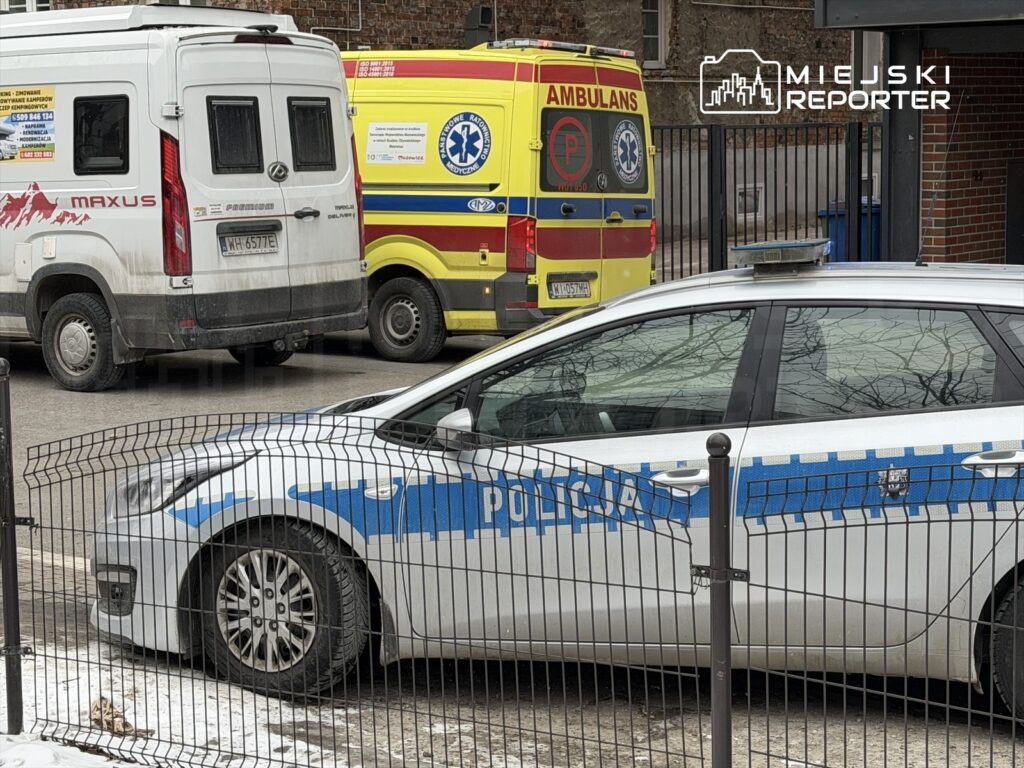 Radiowóz policyjny zaparkowany na ulicy obok ambulansu oraz białego pojazdu dostawczego, z ogrodzeniem w tle.