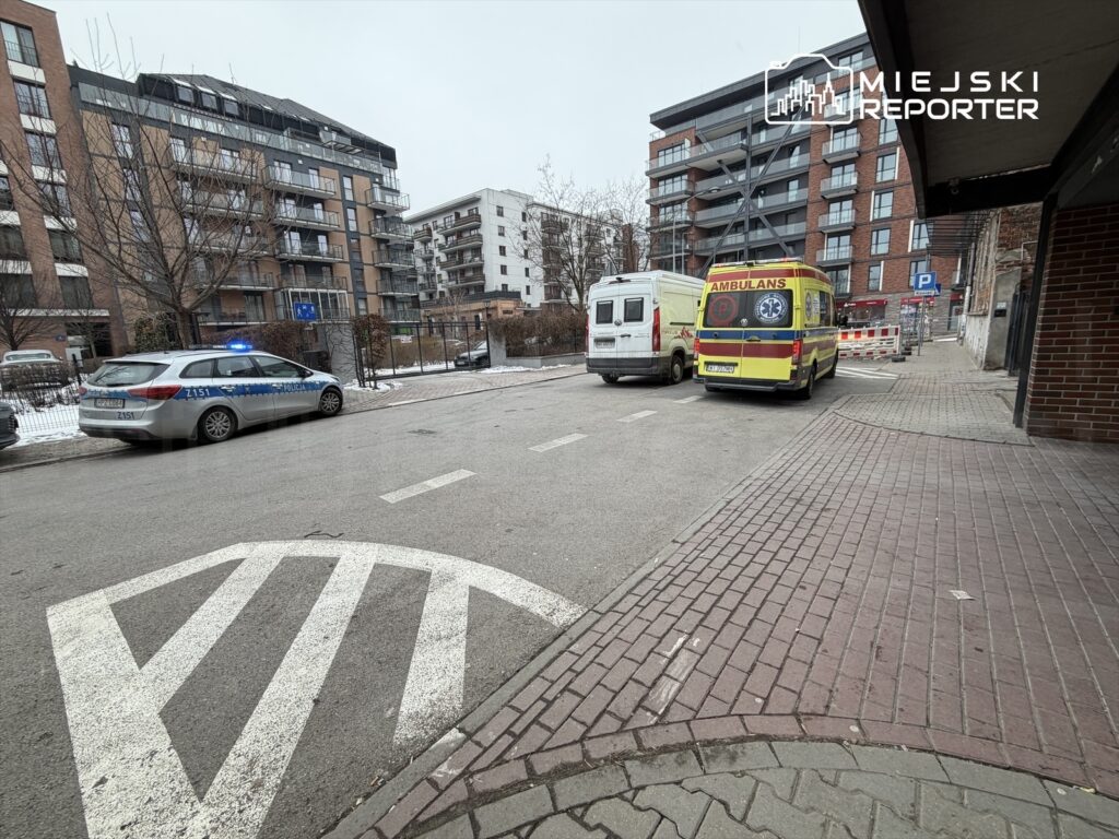 Radiowóz policyjny z włączonymi sygnałami świetlnymi oraz karetka pogotowia zaparkowane na ulicy w miejskim otoczeniu.