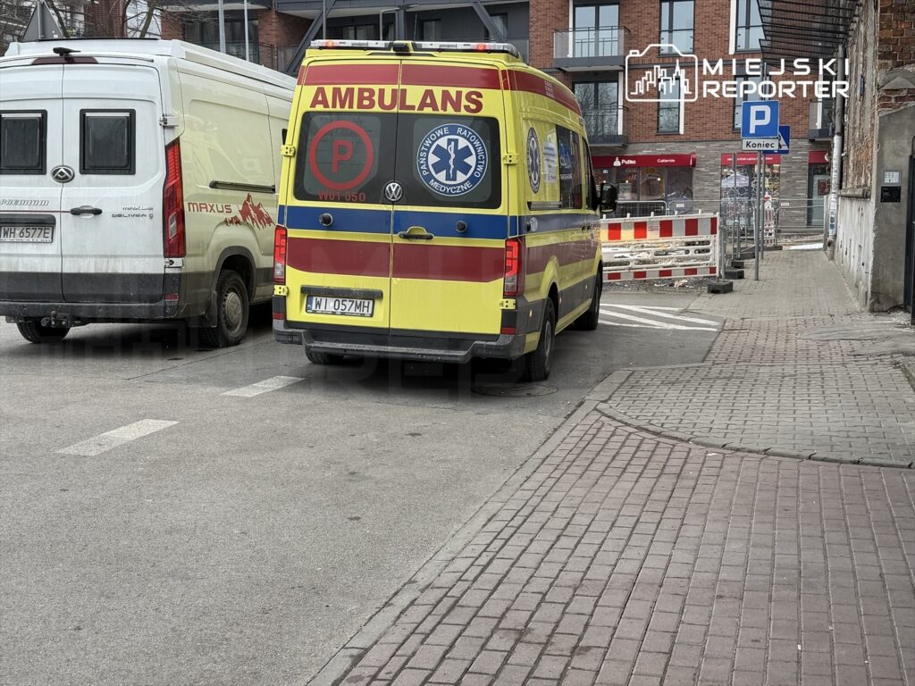 Karetka pogotowia zaparkowana na ulicy obok białego vana, w tle widać prace budowlane.