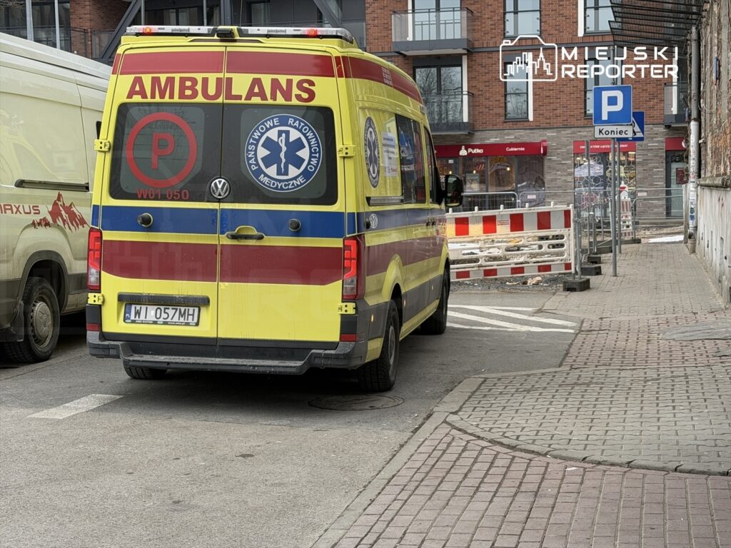 Karetka pogotowia stojąca przy wąskiej ulicy, obok zaparkowanego samochodu dostawczego, z oznaczeniem „AMBULANS”.