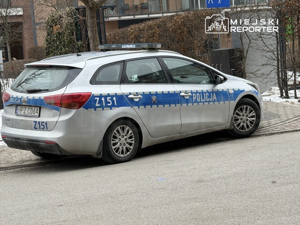 Policyjny samochód zaparkowany na chodniku przy nowoczesnym budynku mieszkalnym w zimowym otoczeniu.