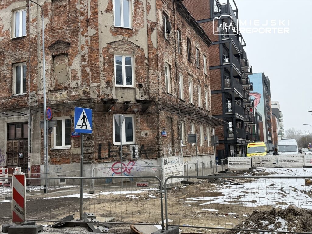 Zniszczona kamienica z odsłoniętą cegłą oraz nowoczesny budynek mieszkalny w tle, w otoczeniu placu budowy.