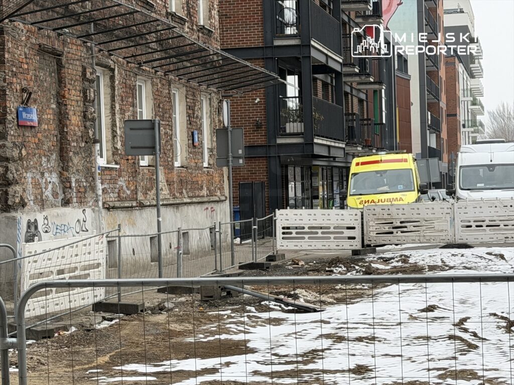 Na ulicy z widocznymi barierkami budowlanymi stoi żółta karetka pogotowia, obok białego vana.
