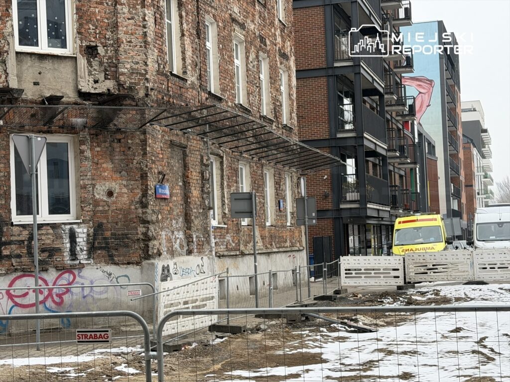 Zniszczona elewacja budynku z widocznymi szczelinami i graffiti, obok nowoczesne budynki mieszkalne oraz pojazdy na placu budowy.
