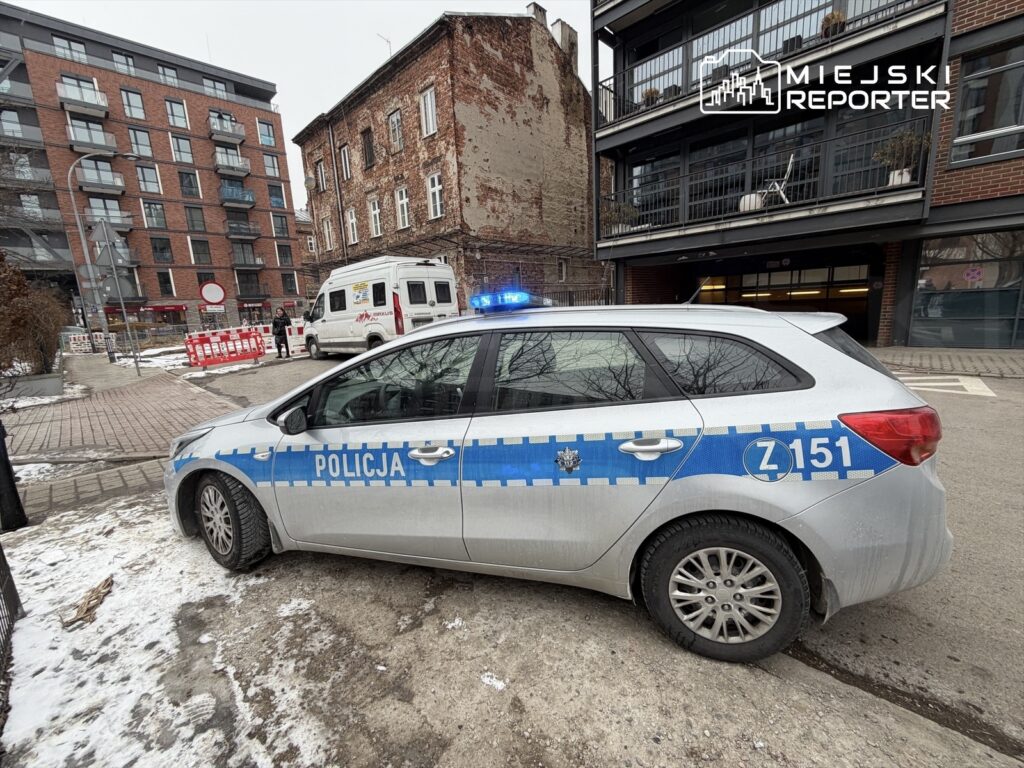 Policyjny radiowóz z włączonymi sygnałami świetlnymi zaparkowany na ulicy obok nowoczesnego budynku mieszkalnego.