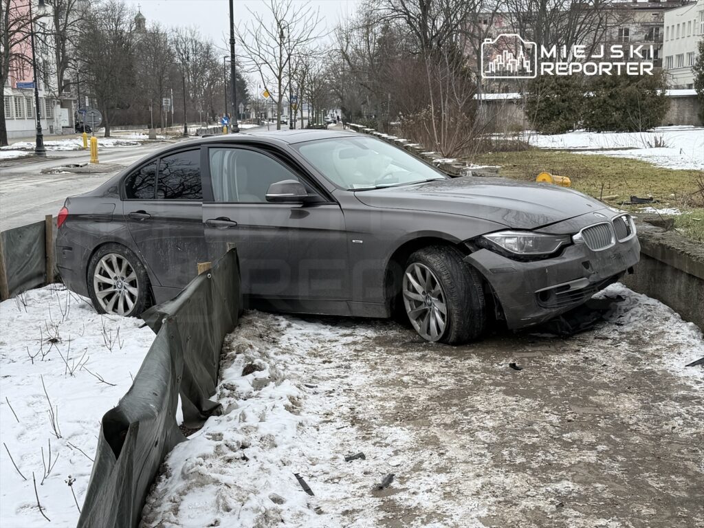 Samochód osobowy marki BMW utknął na krawężniku w otoczeniu śniegu i ogrodzenia z materiału.