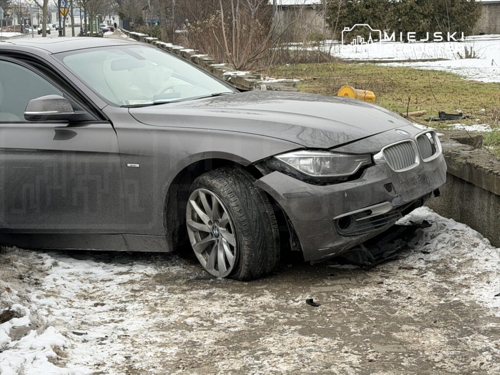 Uszkodzony samochód marki BMW zaparkowany na poboczu drogi, z jednym z przód kół wbitym w ziemię i widocznymi śladami po wypadku.