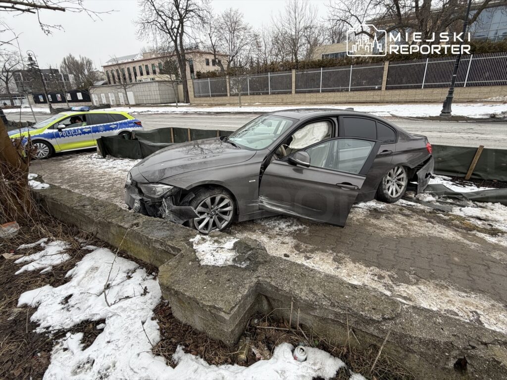 Uszkodzony samochód osobowy zaparkowany na chodniku obok radiowozu policji w zimowej scenerii.