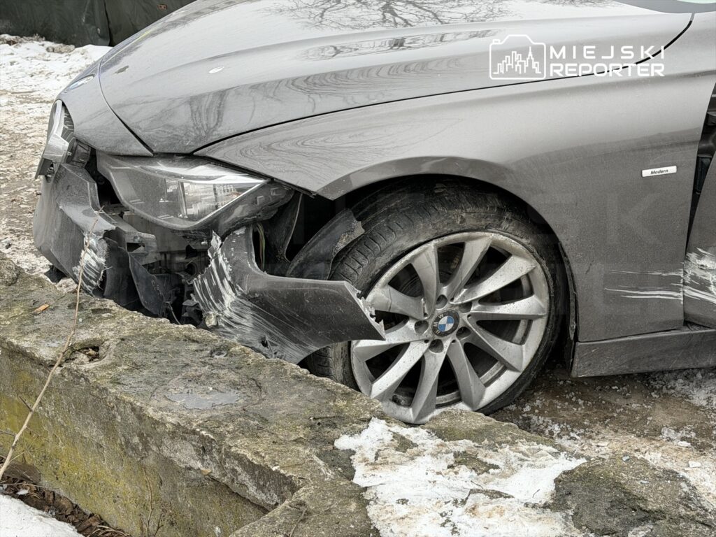 Uszkodzony samochód osobowy marki BMW z wygiętym przodem stoi na ośnieżonym poboczu obok kamiennego muru.