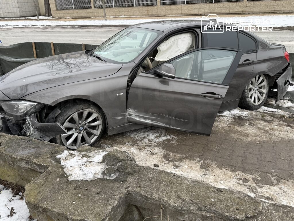 Samochód osobowy w kolorze szarym z uszkodzoną przednią częścią stoi na chodniku, obok widoczny jest kamienny mur i śnieg.