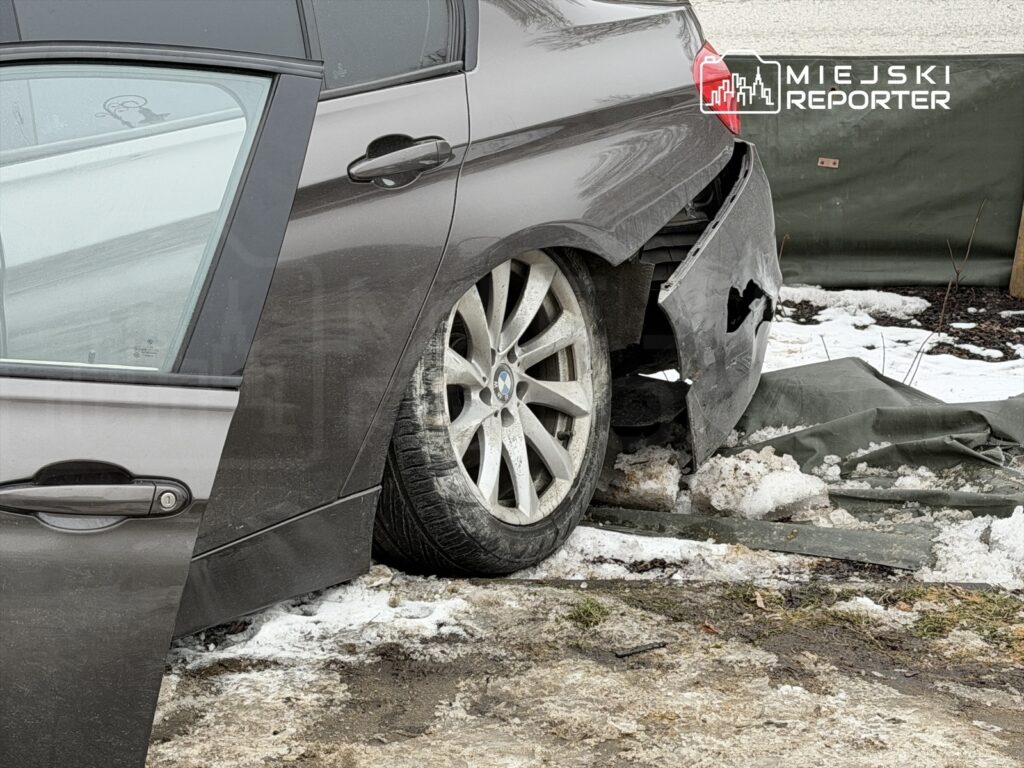 Uszkodzony samochód osobowy z otwartymi drzwiami, stojący na śniegu obok zniszczonego płotu.