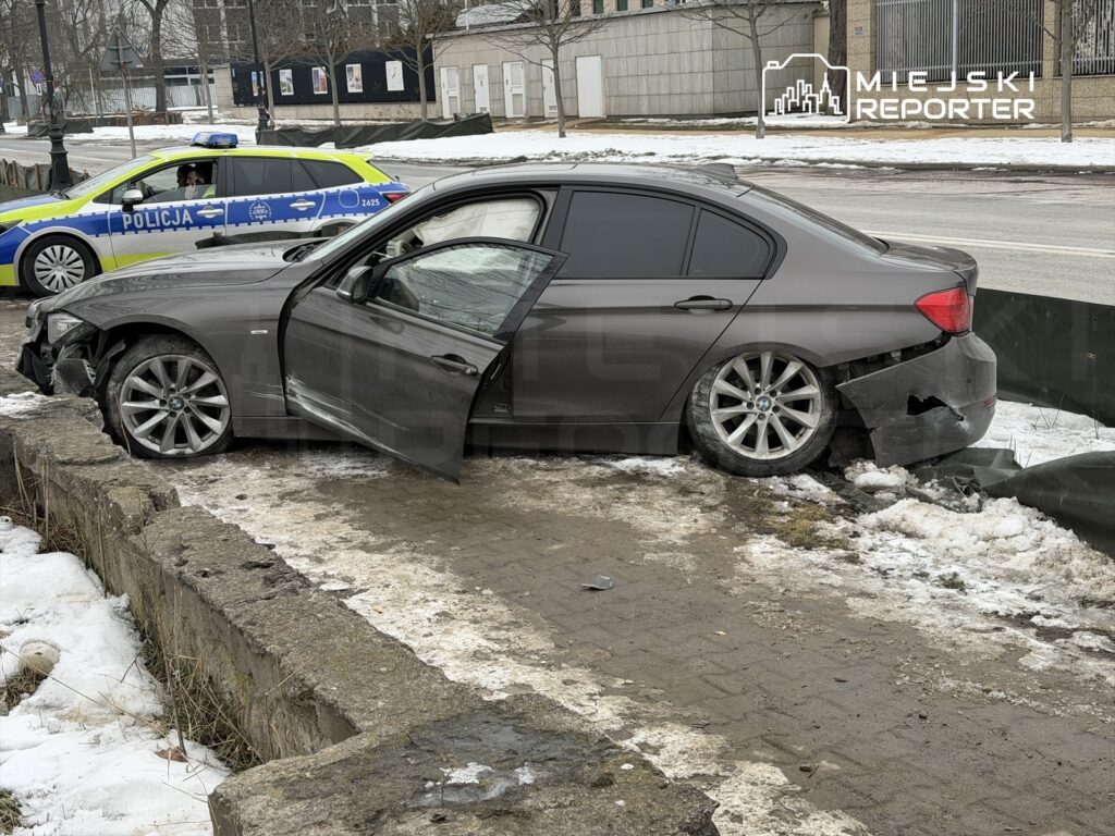 Uszkodzony samochód osobowy stoi na krawężniku, z otwartymi drzwiami, obok patrolu policji. W tle widać zaspy śniegu.