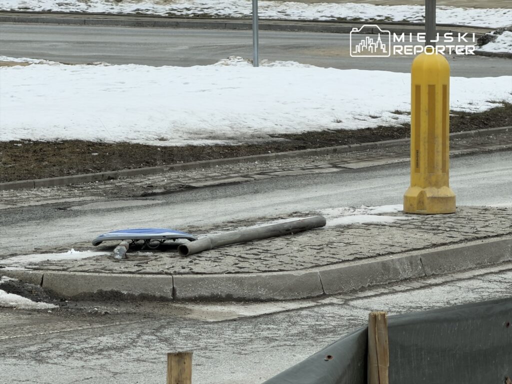 Złamany znak drogowy leży na chodniku obok żółtej słupka, w otoczeniu śniegu i nieutwardzonej nawierzchni.