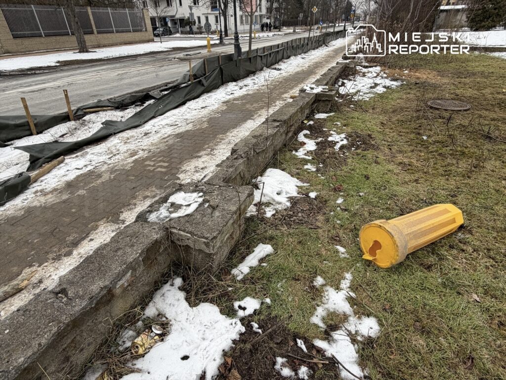 Zniszczony, żółty pojemnik leży na trawniku obok chodnika otoczonego śniegiem i prowizorycznym ogrodzeniem.