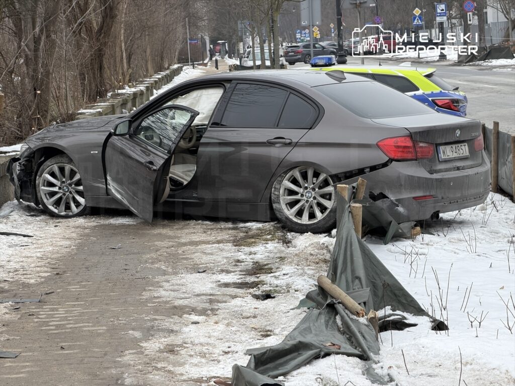 Samochód osobowy BMW z otwartymi drzwiami stoi na poboczu drogi, obok uszkodzonego ogrodzenia w zimowej scenerii.