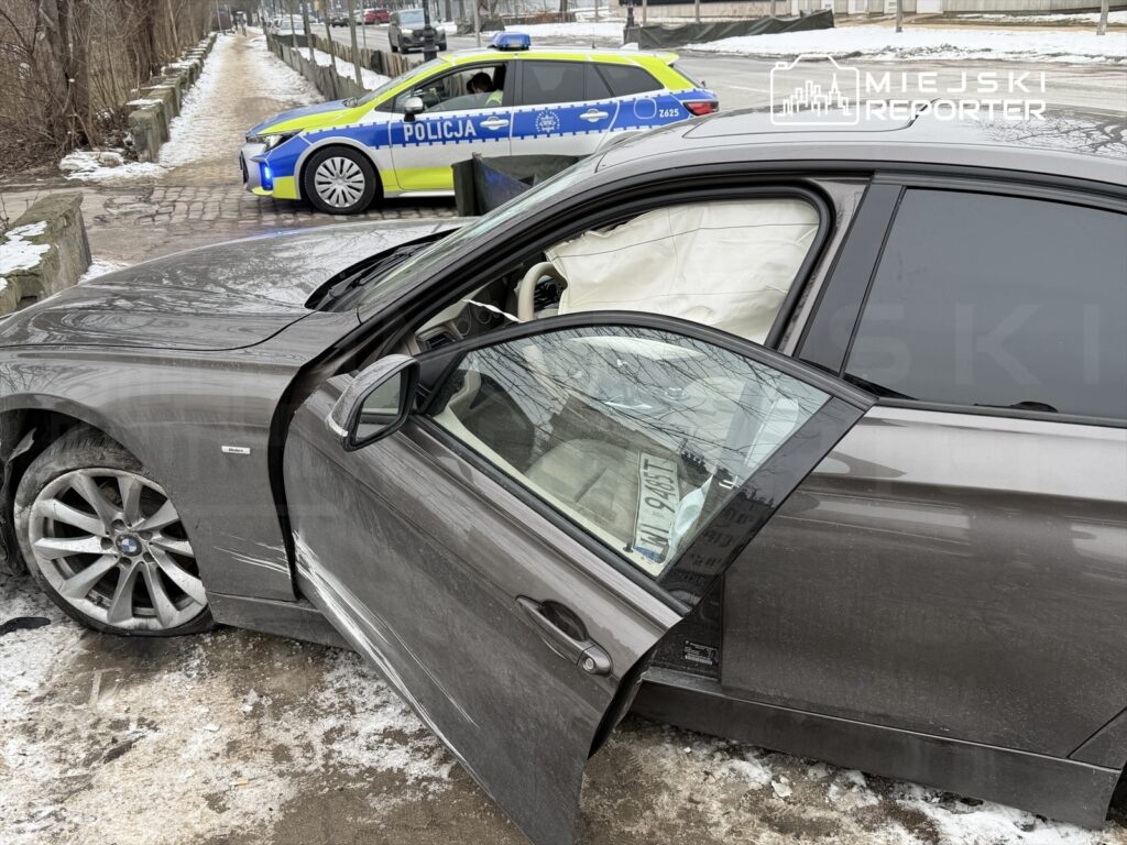 Uszkodzony samochód osobowy stoi na chodniku obok policyjnego radiowozu, w tle widać zamarzniętą nawierzchnię.