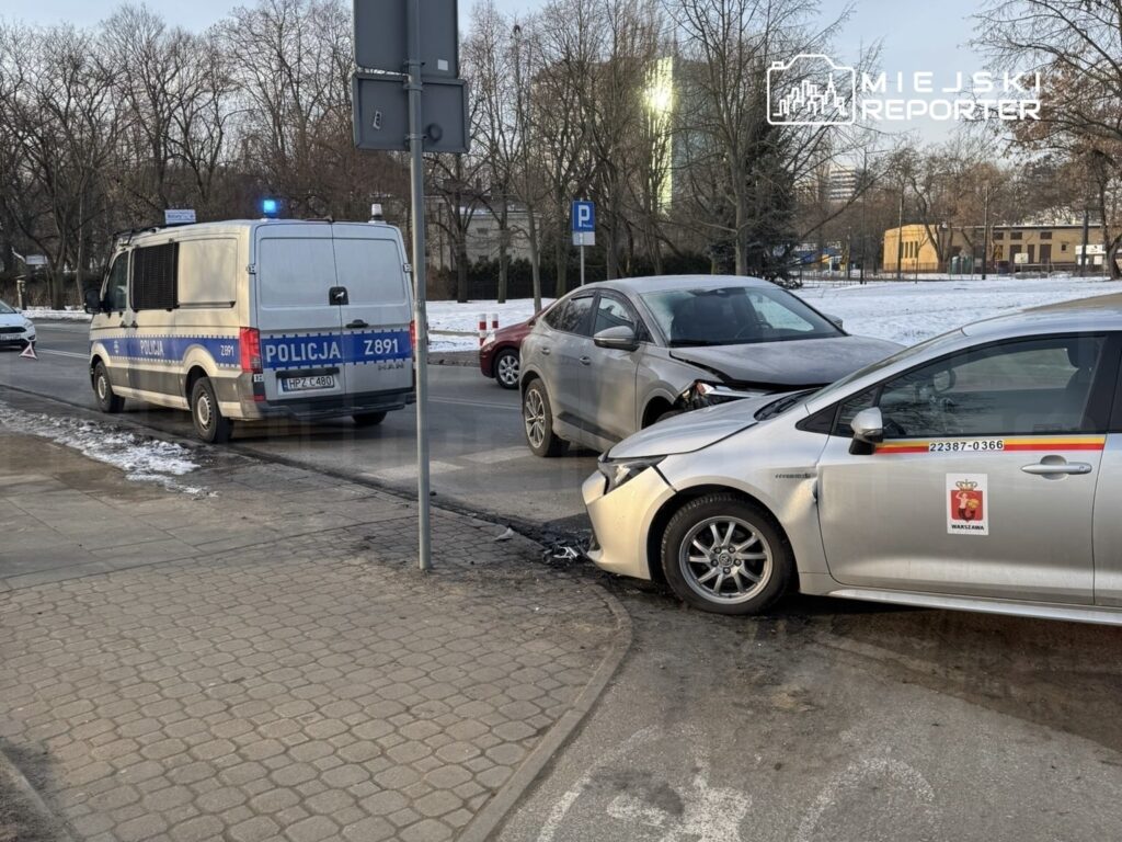Dwa uszkodzone samochody osobowe stoją na drodze, obok policyjny radiowóz z włączonymi sygnałami świetlnymi.