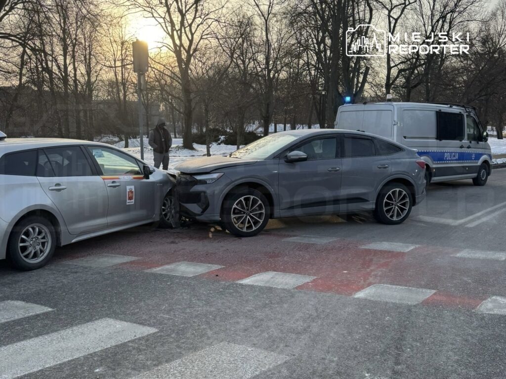 Dwa uszkodzone samochody osobowe na przejściu dla pieszych, obok stoi radiowóz policji. W tle widoczny mężczyzna.