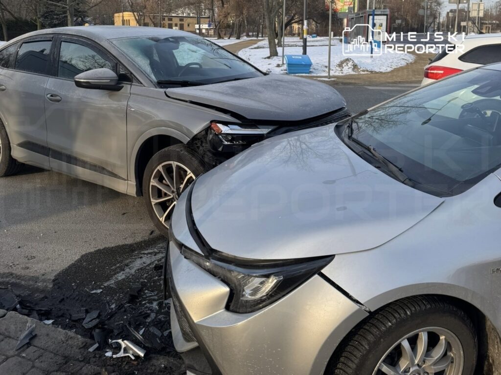 Dwa uszkodzone samochody osobowe stoją na ulicy, jeden z nich ma zgnieciony przód, a na jezdni leżą fragmenty plastików.