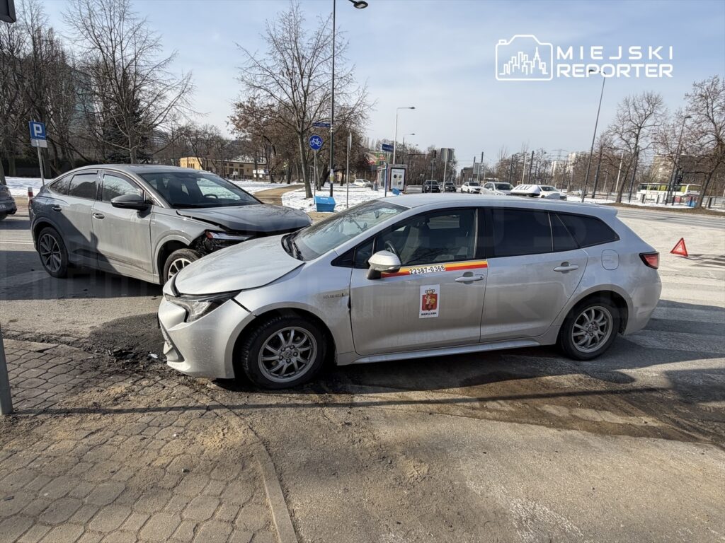 Dwa uszkodzone samochody osobowe na skrzyżowaniu, w tle widoczne znaki drogowe i parkowane pojazdy.