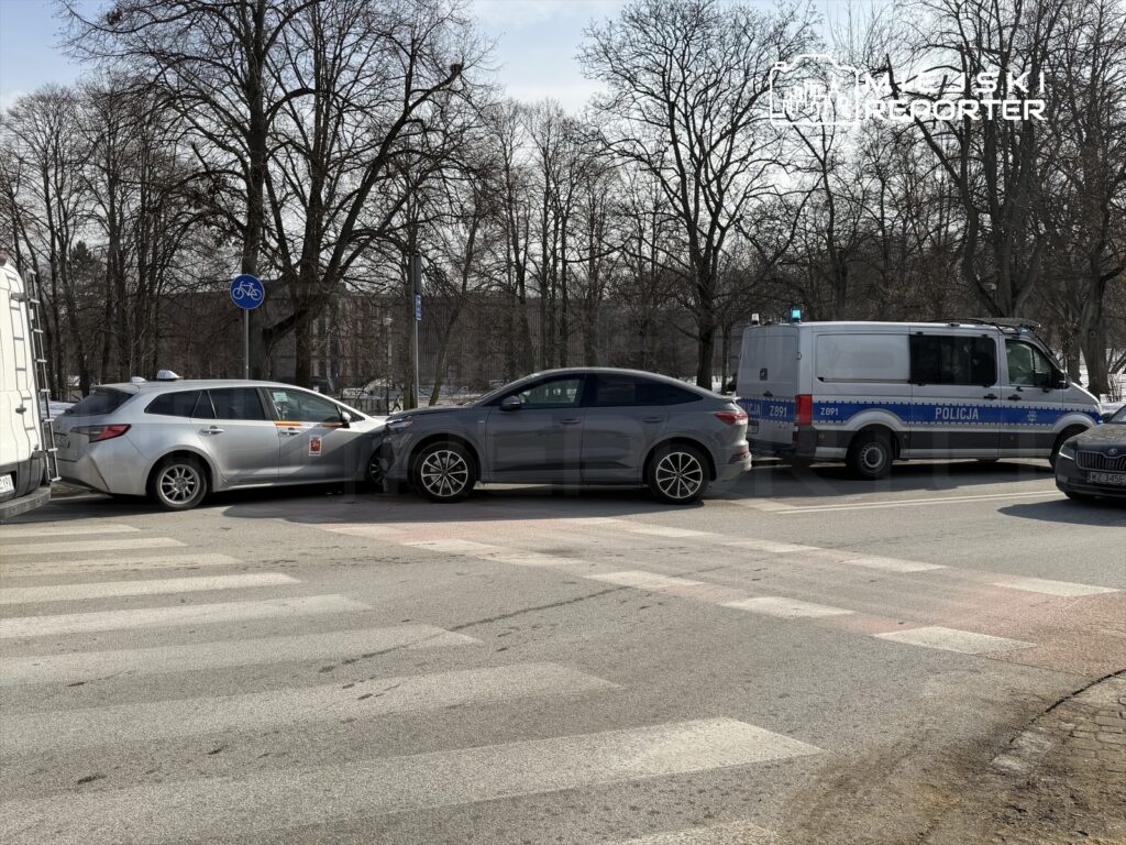 Dwa uszkodzone samochody osobowe zderzone na przejściu dla pieszych, obok radiowóz policyjny z włączonymi sygnałami.