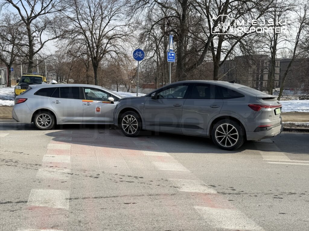 Dwa samochody osobowe zderzone na skrzyżowaniu, przy chodniku otoczonym drzewami.