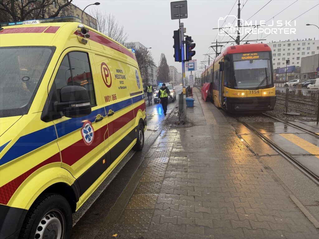 Ambulans z włączonymi sygnałami świetlnymi stoi obok przystanku, obok zatrzymanego tramwaju. Na chodniku znajdują się policjanci.