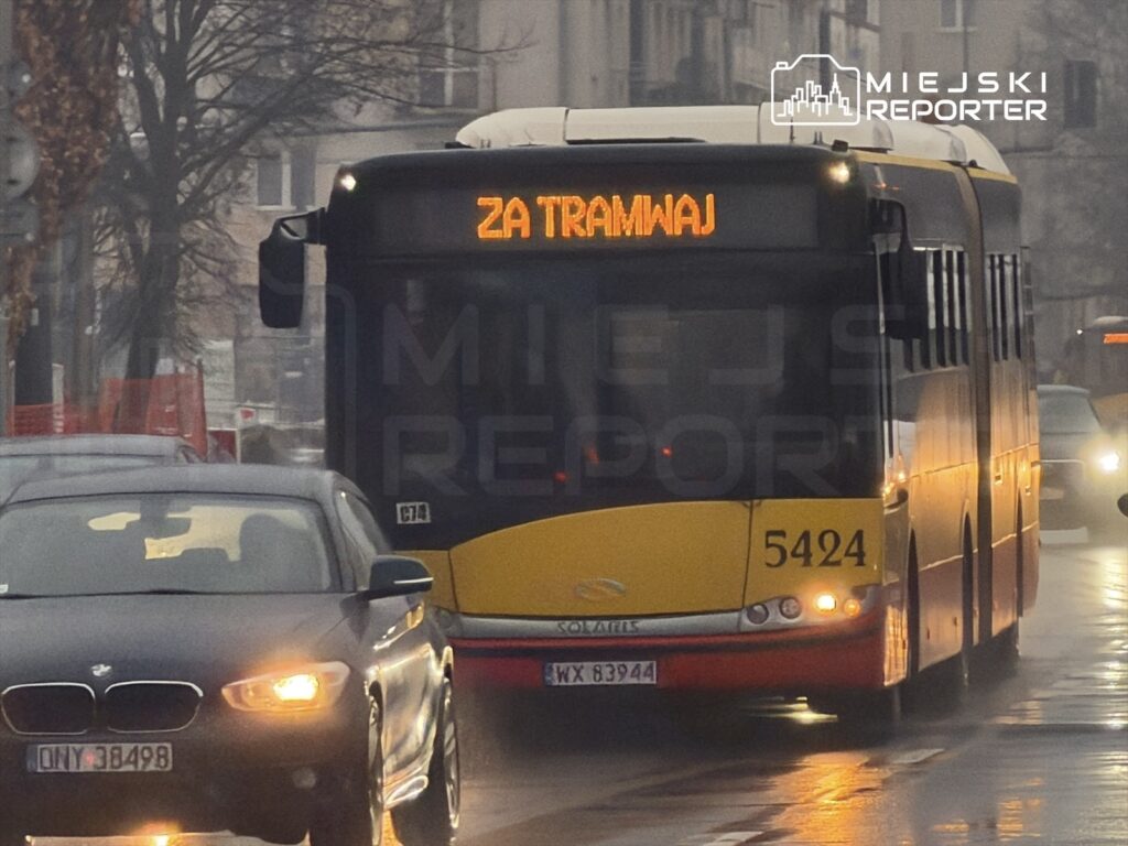 Autobus z wyświetlaczem "ZA TRAMWAJ" porusza się po deszczowej ulicy, mijając inne pojazdy.