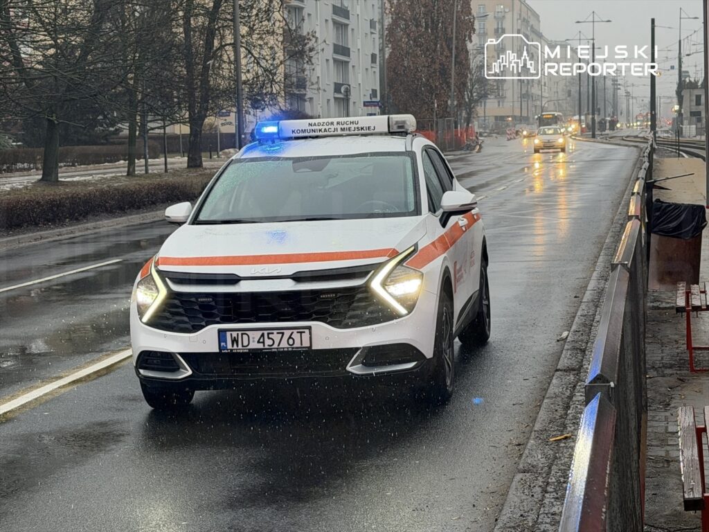 Samochód służbowy z niebieskim włączonym sygnałem na deszczu, poruszający się po miejskiej ulicy.