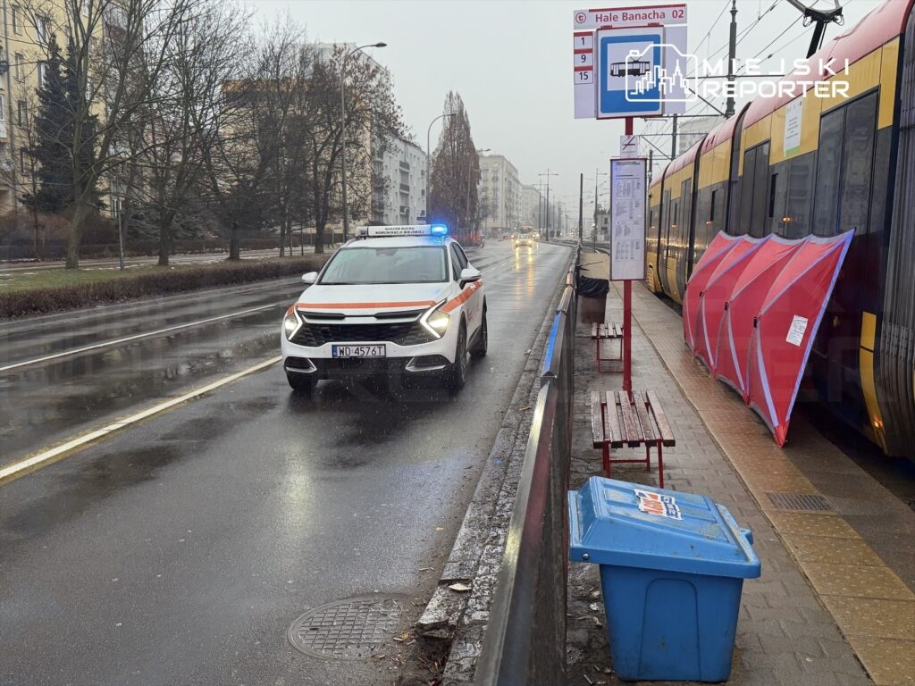 Radiowóz policyjny z włączonymi sygnałami świetlnymi stoi na ulicy obok przystanku tramwajowego w deszczowy dzień.