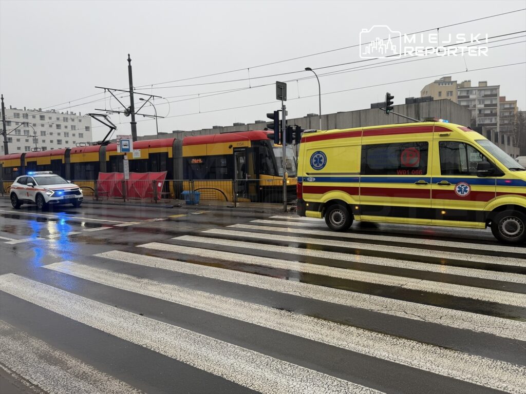 Policyjny radiowóz i karetka stoją na pasach, obok zatrzymanego tramwaju w deszczowym otoczeniu.