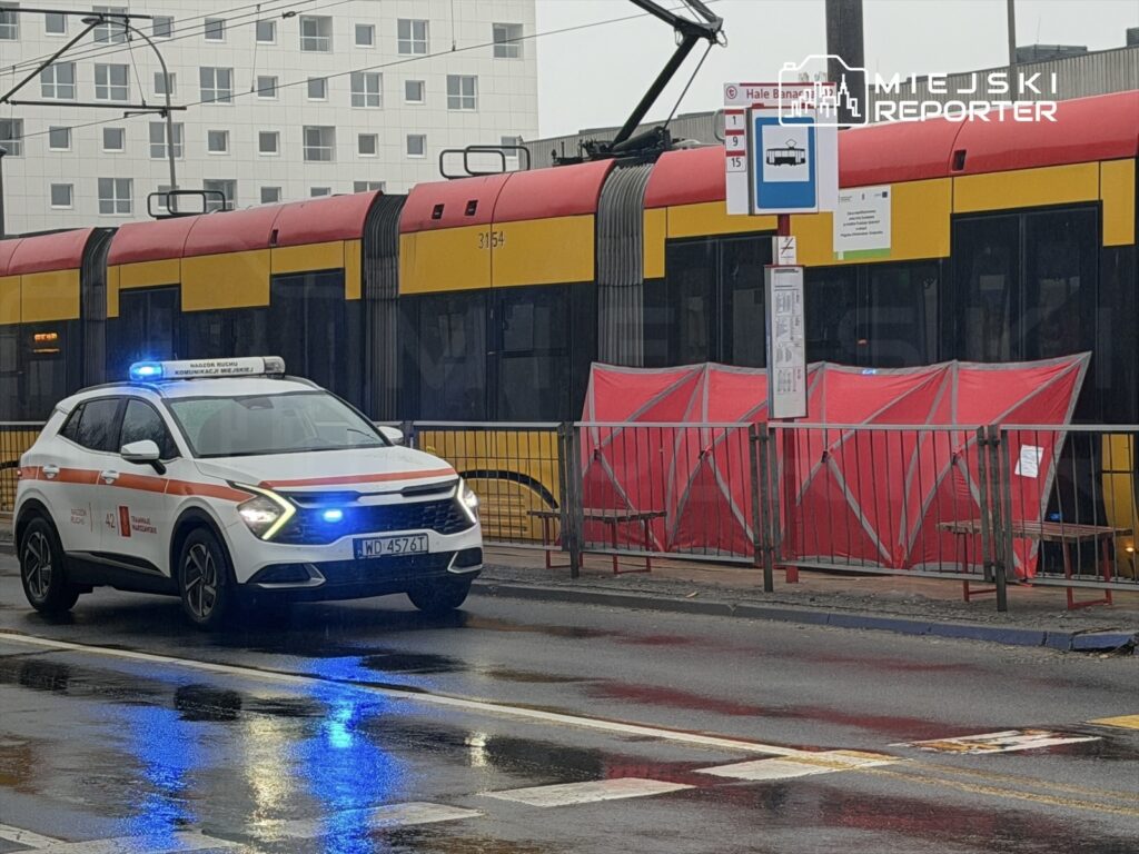 Pojazd służbowy z włączonymi sygnałami świetlnymi stoi obok tramwaju na przystanku, w otoczeniu deszczowej ulicy.