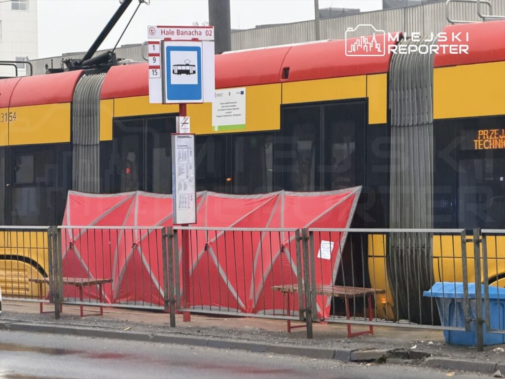 Tramwaj w kolorze czerwono-żółtym zatrzymany przy przystanku, obok czerwony namiot i tablica z rozkładem.