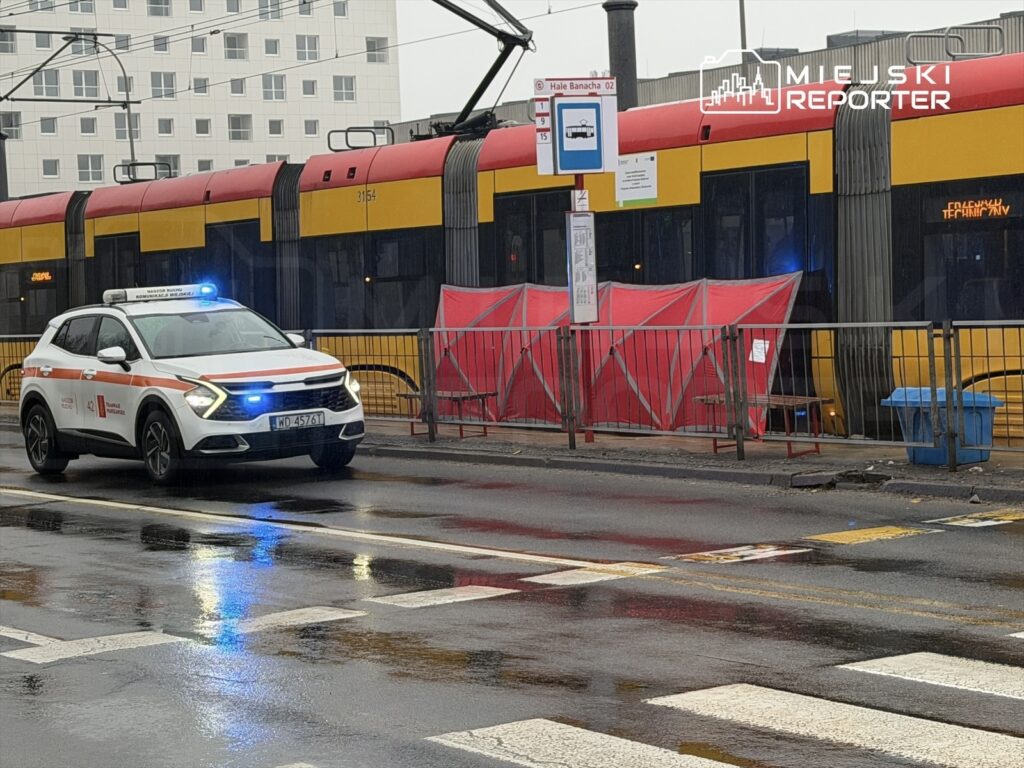 Radiowóz z włączonymi sygnałami świetlnymi stoi obok przystanku tramwajowego, obok zatrzymanego tramwaju.