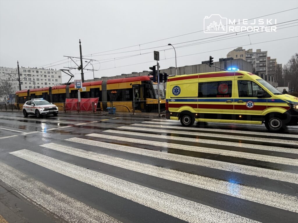Karetka pogotowia z włączonymi sygnałami świetlnymi stoi na pasach, obok zatrzymanego tramwaju w deszczowej scenerii.
