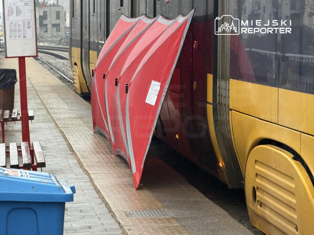 Czerwony namiot ochronny przylegający do drzwi tramwaju na przystanku, w tle widoczna peronowa tablica informacyjna.