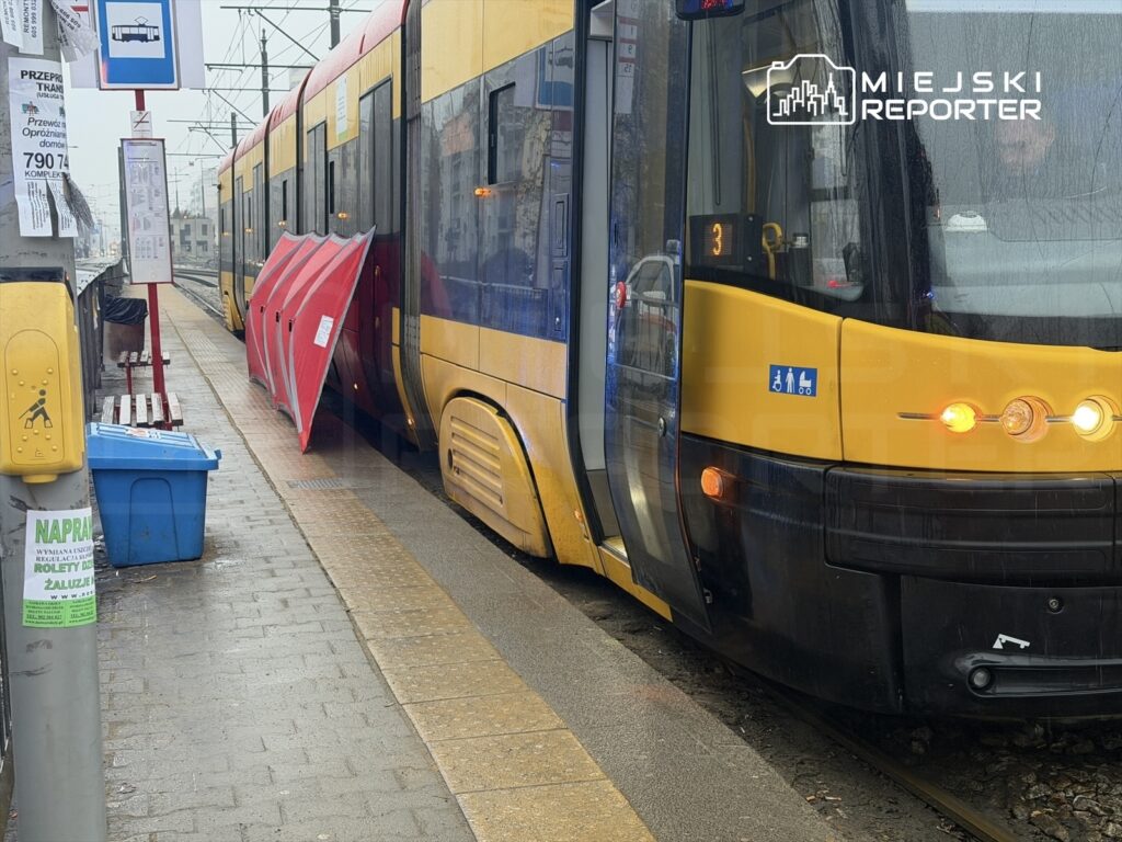 Tramwaj zatrzymany na przystanku, z otwartymi drzwiami i czerwonym zabezpieczeniem, w deszczowej aurze. Na tle widoczne są ławki i znak przystanku.
