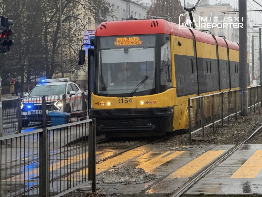 Tramwaj w kolorach żółtym i czerwonym zatrzymany na przystanku obok policyjnego radiowozu z włączonymi sygnałami świetlnymi.