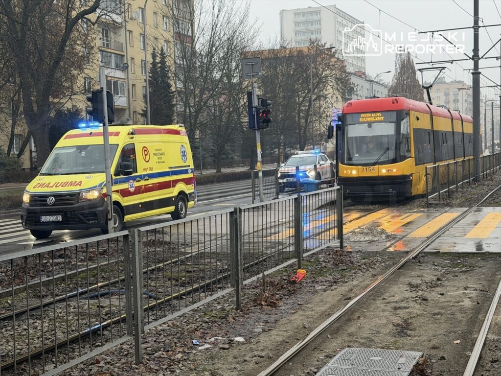 Ambulans z włączonymi sygnałami świetlnymi przejeżdża obok tramwaju na ulicy w deszczowy dzień.