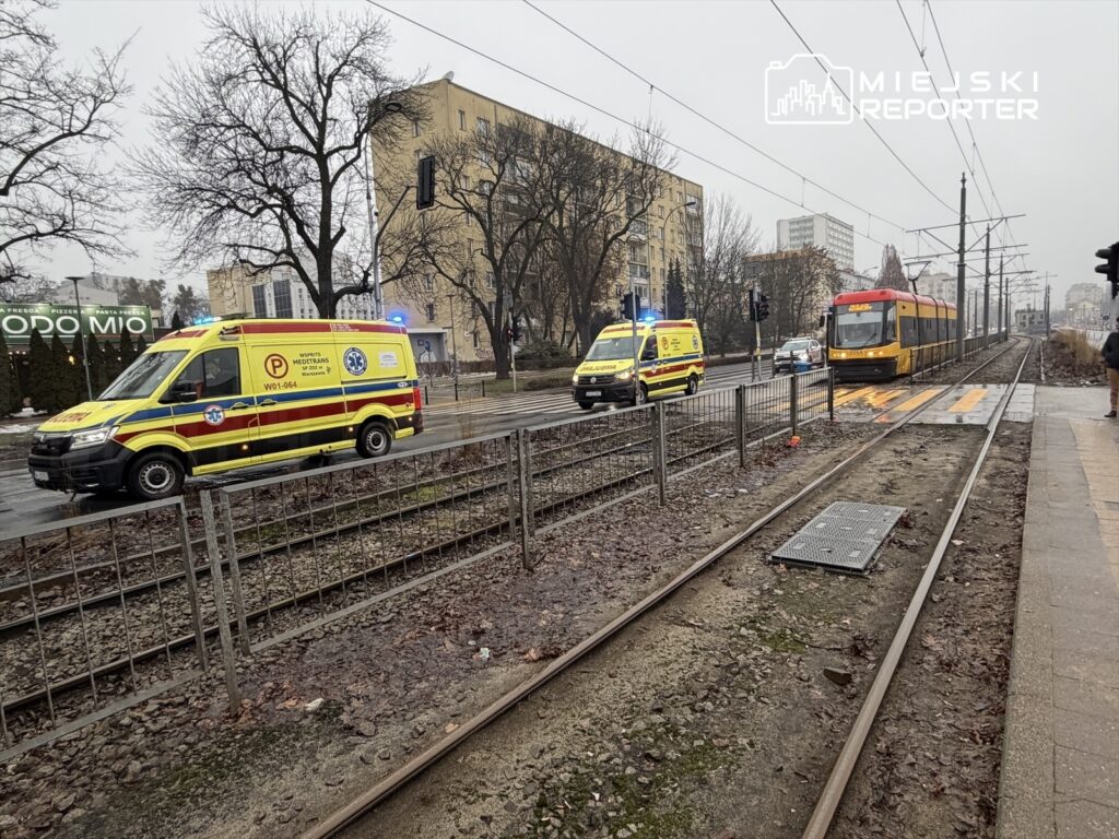 Dwa karetki pogotowia z włączonymi sygnalami świetlnymi przejeżdżają obok tramwaju na deszczowej ulicy.