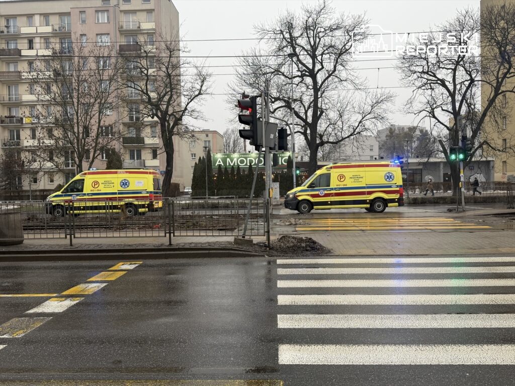 Dwa karetki pogotowia stoją na skrzyżowaniu w deszczowy dzień, obok sygnalizacji świetlnej i budynków mieszkalnych.